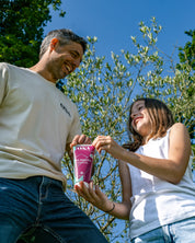 Tube de gel douche parfum verveine tenu par un homme et une fille souriant l'un à l'autre dans un environnement extérieur.
