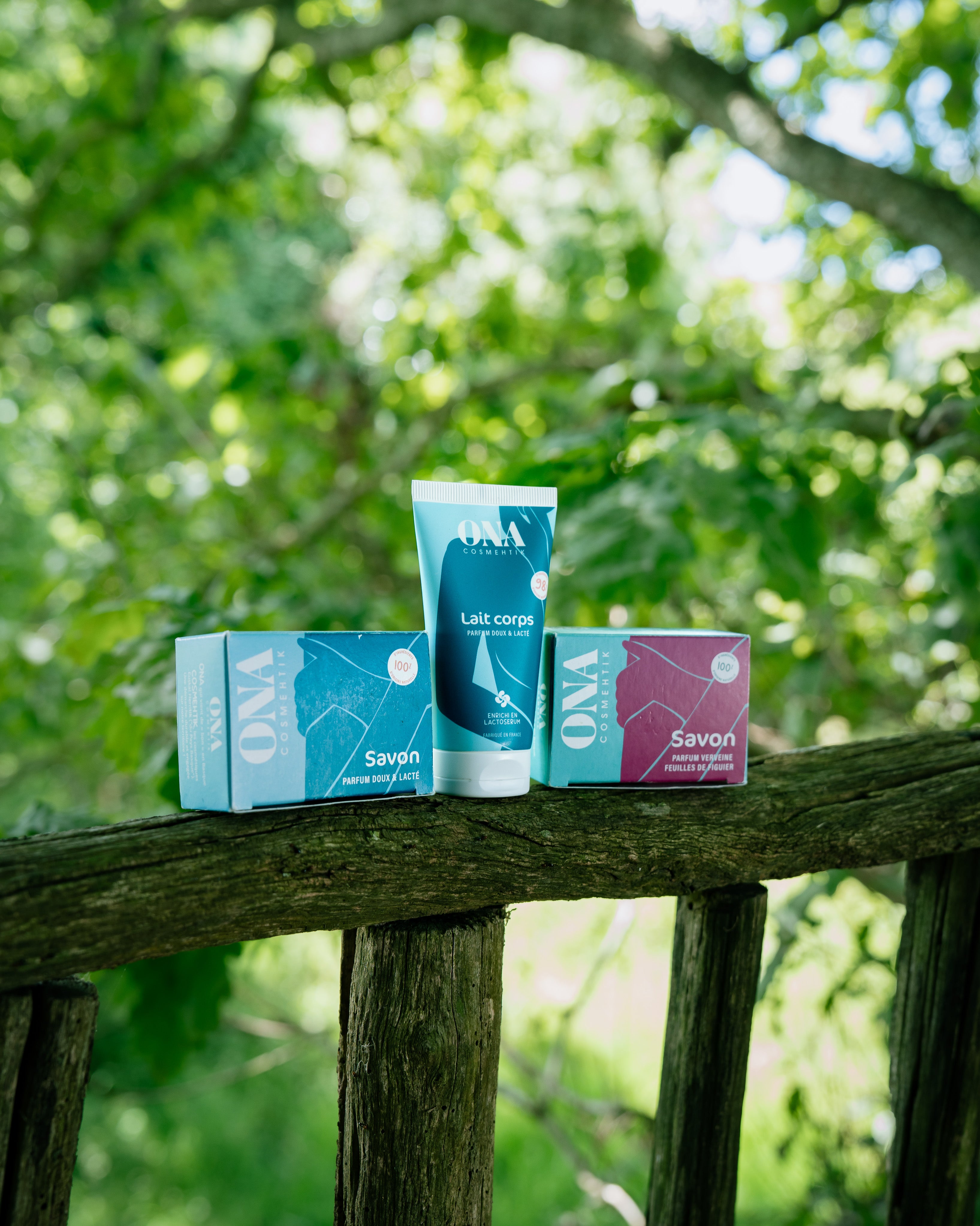 Deux savons et un lait corps de la marque ONA COSMEHTIK posés sur une rambarde en bois en pleine nature. 