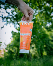 Une main tenant vers le bas un tube de shampoing, avec en arrière-plan des arbres et de la verdure. 