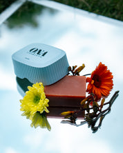 Porte savon et savon de la marque ONA COSMEHTIK entourés de fleurs jaunes et oranges. 