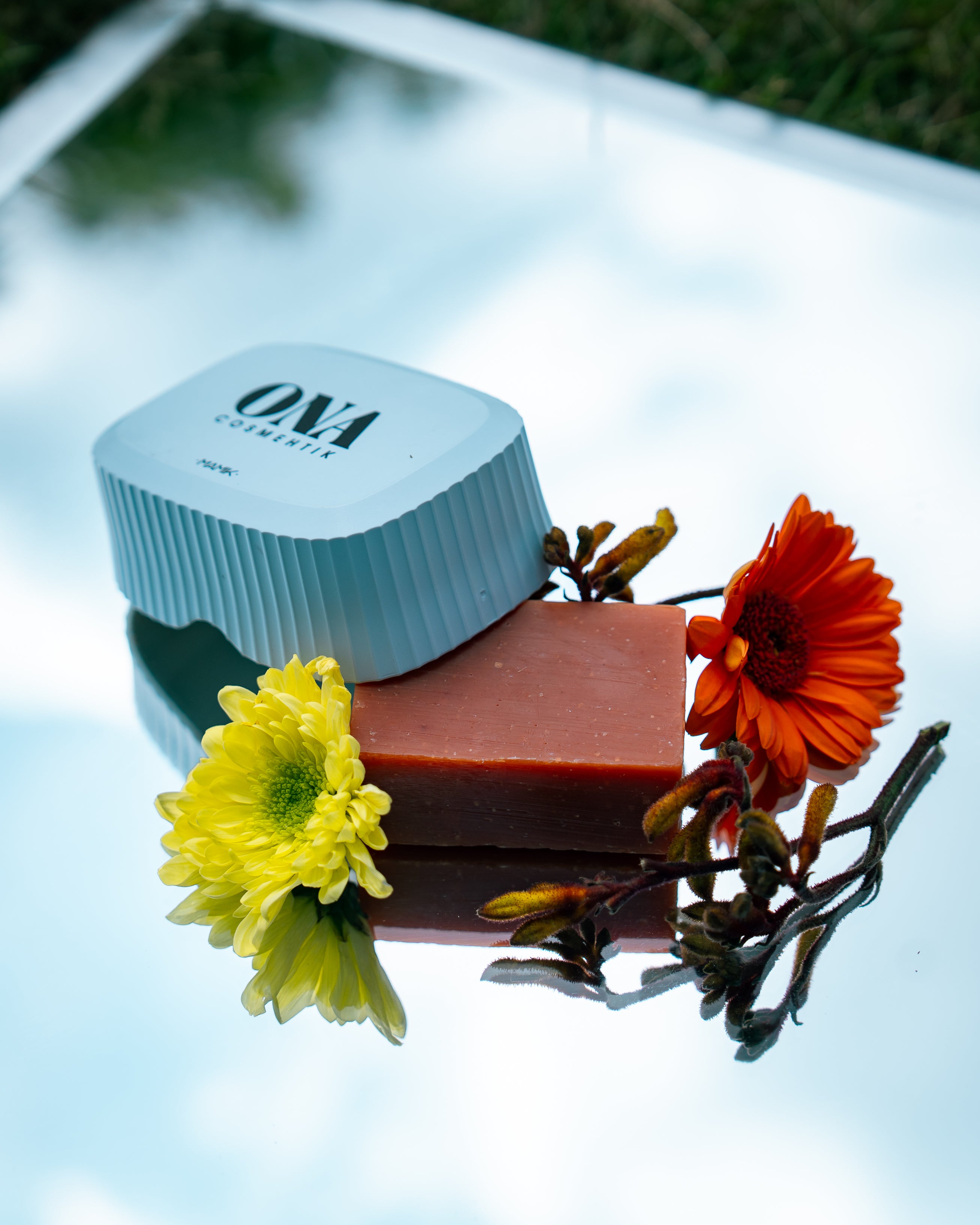Porte savon et savon de la marque ONA COSMEHTIK entourés de fleurs jaunes et oranges. 