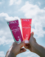 Deux tubes de gel douche de la marque ONA COSMEHTIK tenus par deux mains avec le ciel et les nuages en fond. 