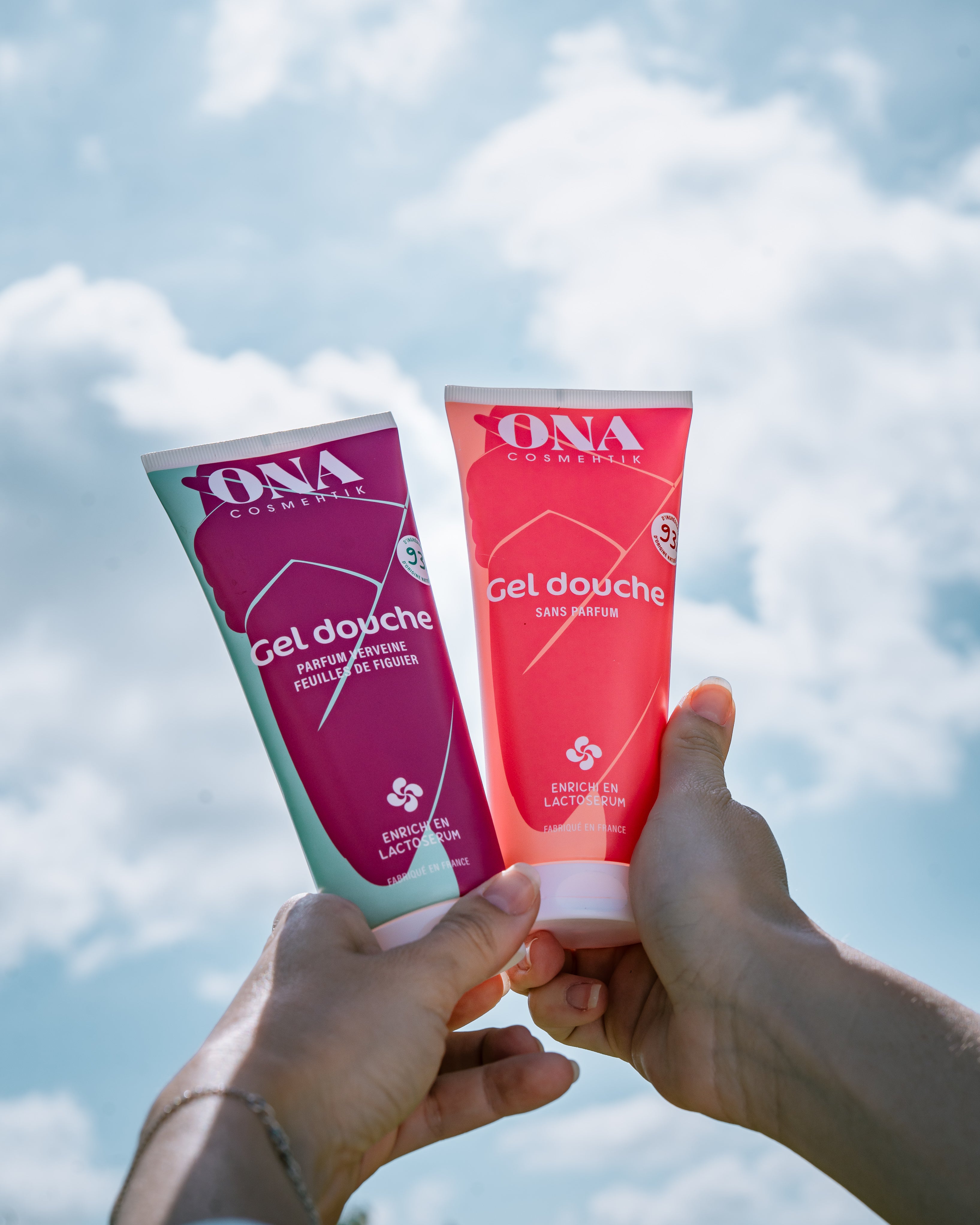 Deux tubes de gel douche de la marque ONA COSMEHTIK tenus par deux mains avec le ciel et les nuages en fond. 